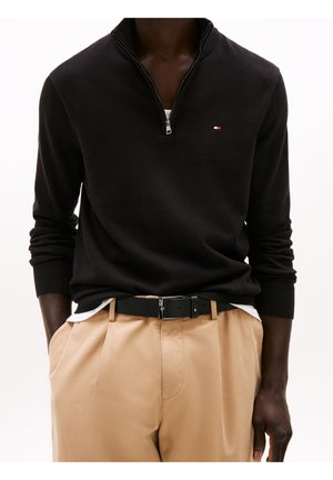 Uomo che indossa maglione nero con zip a quarto e piccolo logo, pantaloni beige, cintura nera con logo, mani in tasca, su sfondo bianco uniforme.