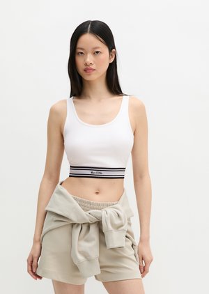 Femeie care poartă un crop top alb cu bandă neagră în dungi și pantaloni scurți bej, hanorac bej legat în jurul taliei, stând în picioare pe un fundal simplu.