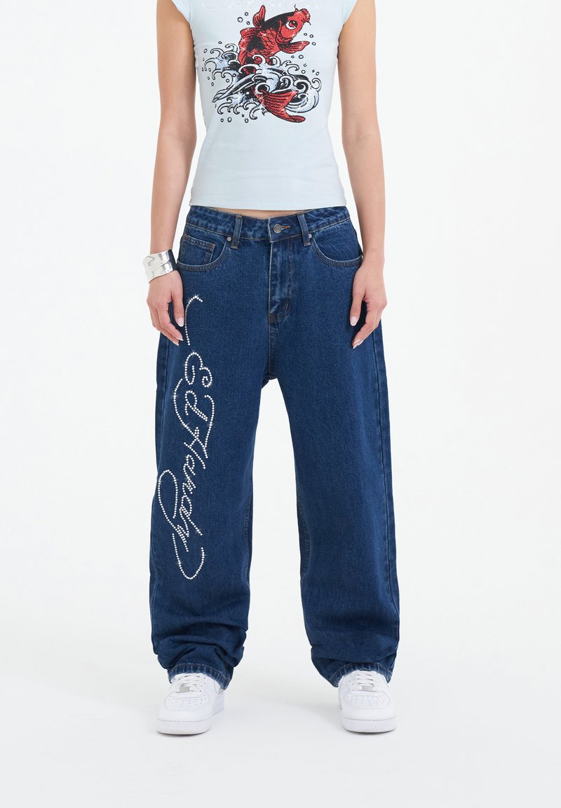Jeans baggy de mezclilla azul con un acabado texturizado, presentan acentos de rhinestones blancos y un diseño de escritura decorativa en el costado. Zapatillas blancas.