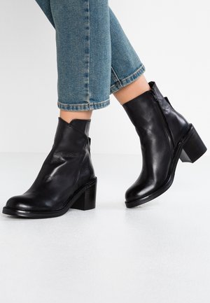 Schwarze knöchelhohe Lederstiefel mit Blockabsätzen, getragen zu blauen, gekürzten Jeans, vor schlichtem Hintergrund.