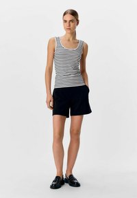 Top sin mangas a rayas en blanco y negro con escote en forma de U, combinado con pantalones cortos negros. La modelo posa con mocasines negros sobre un fondo simple.