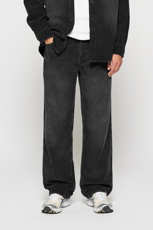 Man wearing black corduroy pants, black corduroy shirt, white sneakers, and white undershirt, standing with one hand in pocket.
