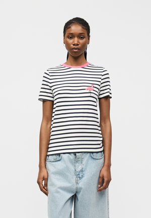 Femeie purtând un tricou cu dungi alb-negru, cu o aplicație cu un flamingo roz și jeansi din denim bleu deschis, stând în fața unui fundal alb.