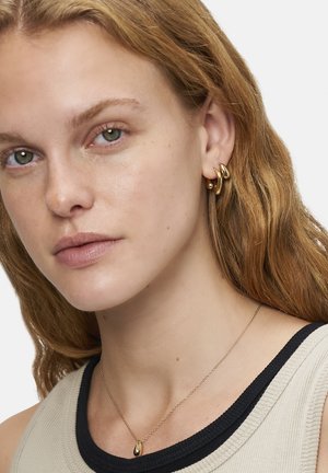Goldene Creolen mit einem glatten, geschwungenen Design und einer Tropfenanhänger-Halskette. Das Model hat welliges Haar und trägt ein neutrales Outfit mit Rippstruktur.