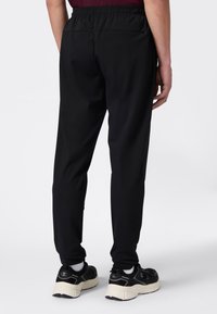 Persoon in zwarte tapse broek, witte sokken en zwarte sneakers met beige zolen, staand tegen een effen lichte achtergrond.