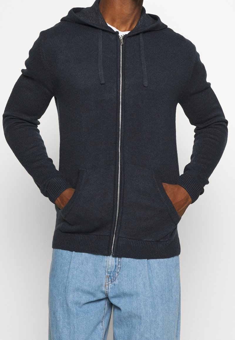 Felpa con zip di un blu scuro realizzata in tessuto testurizzato, dotata di cappuccio, polsini a coste e due tasche laterali con dettagli marroni.