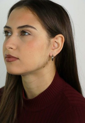 Jonge vrouw met lang donker haar, zilveren oorring in de vorm van een ring en bordeauxrode geribbelde coltrui, kijkt iets naar boven en naar links, neutrale achtergrond.