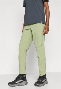 Ανοιχτοπράσινο παντελόνι εξωτερικού χώρου με στενή εφαρμογή, κατασκευασμένο από ομαλό, ελαφρύ ύφασμα. Συνδυασμένο με μαύρα παπούτσια πεζοπορίας με γκρι λεπτομέρειες.