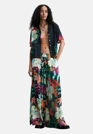 Frau mit lockigem Haar, die ein schwarzes kurzärmeliges Hemd mit Blumenmuster, passende weite Hosen, rosa Bikinioberteil, schwarze Sandalen und eine Herz-Halskette trägt.