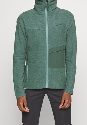Hombre con chaqueta de forro polar verde con cremallera y cuello alto, con bolsillo en el pecho de color verde oscuro, combinado con pantalones negros.