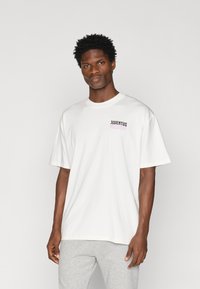 T-shirt oversized branca de algodão com um logótipo preto e rosa "JUVENTUS MILLES8" na área do peito esquerdo. Mangas curtas, decote redondo.