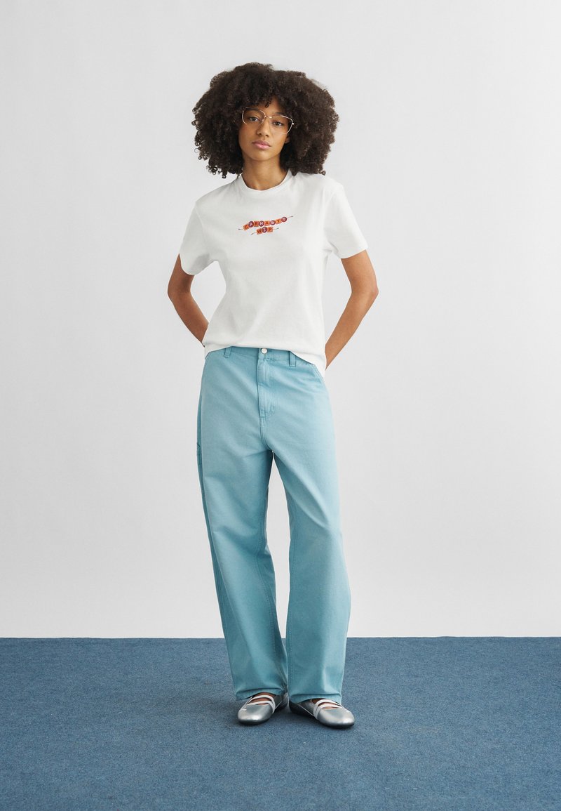 Γυναίκα με σγουρά μαλλιά που φορά γυαλιά, λευκό T-shirt με σχέδιο, ανοιχτό μπλε φαρδιά παντελόνια και ασημένια μπαλαρίνες, στέκεται σε μπλε χαλί.