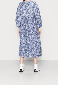 Osoba oblečená v modrých květinových šatech midi s dlouhým rukávem a bílých teniskách na vysoké podrážce, stojící zády k fotoaparátu na jednotném pozadí.