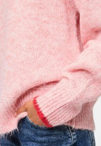 Miękki różowy puszysty sweter z czerwonym wykończeniem na mankietach, noszony z ręką w kieszeni niebieskich dżinsów.