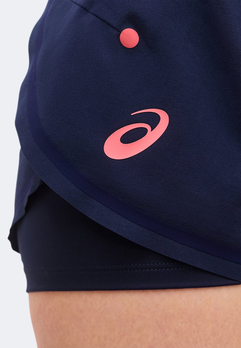 Nahaufnahme von marineblauen Sportshorts mit überlappendem Saum und pinkem Logo sowie Knopfdetail an der Seite oberhalb des nackten Oberschenkels.
