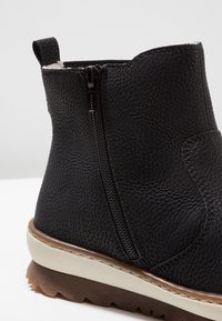 Svart läderankelboot med en strukturerad yta, sidodragkedja och en lätt vit och brun sula. Har en bakre dragflik för enkel påtagning.