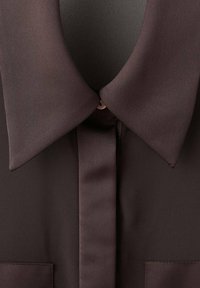 Brown blouse with a pointed collar, smooth satin texture, buttoned placket, and two chest pockets. Minimalist design with a sleek finish.
