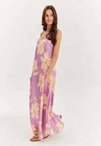 Maxi šaty z jemně fialové látky s velkými krémovými květinovými vzory. Špagetové ramínka a volný silueta. Hnědé sandály.