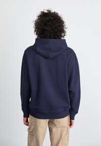 Hoodie bleu marine, tissu en mélange de coton, avec une capuche à cordon de serrage et des poignets et un ourlet côtelés. Porté avec un pantalon cargo beige.