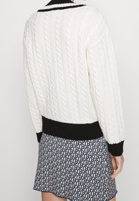 Bílý svetr s kabelovým vzorem, černým žebrovaným límcem a lemem, doplněný vzorovanou sukní s malými tmavě modrými písmeny.