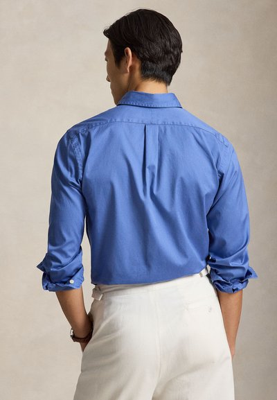 Homme vu de dos portant une chemise bleue vif à manches longues retroussées et un pantalon blanc sur un fond uni.