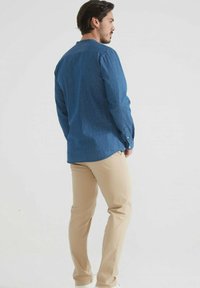 Homme debout, portant une chemise en denim bleu à manches longues et un pantalon beige, tourné sur le côté devant un fond clair.