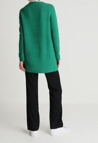 Grünes, langärmliges Strickoberteil mit lockerem Sitz, kombiniert mit schwarzen Hosen und weißen Turnschuhen. Glatte Textur und schlichtes Design.