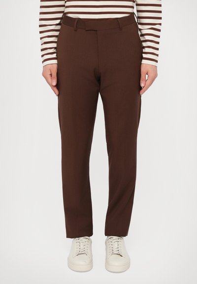 Person, der braune, maßgeschneiderte Hosen, ein langärmliges beige-braun gestreiftes Oberteil und weiße Sneakers trägt, steht vor einem einfarbigen Hintergrund.