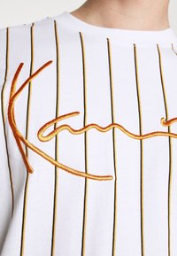 Camisa blanca de manga corta con rayas verticales en negro y amarillo, con texto bordado en naranja en un diseño ondulado.