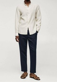 Kangaspaita, vaalea beige, mandariinikauluksella; yhdistettynä tummansinisiin housuihin ja ruskeisiin sandaaleihin, joissa on useita hihnoja.