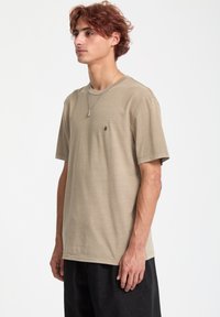 Beige bomulds T-shirt med rund halsudskæring og korte ærmer, med et lille sort logo på venstre side. Subtil tekstur og afslappet pasform.
