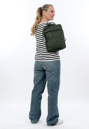 Vrouw die lacht, gekleed in een gestreept shirt en spijkerbroek, met een donkergroene leren rugzak over haar schouder tegen een witte achtergrond.