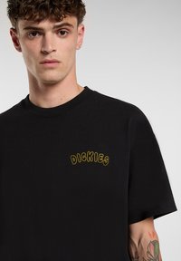 Young man with curly hair wearing a black Dickies t-shirt, showing a colorful tattoo on his forearm against a plain background.