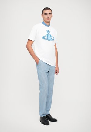 White t-shirt with a blue graphic design, light blue striped trousers, and black shoes. Model stands with one hand in pocket against a white background.