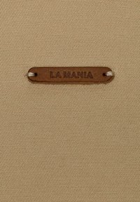 Hnedá kožená etiketa s nápisom "LA MANIA" prišitá na béžovej textúrovanej tkanine s dvoma bielymi stehmi.