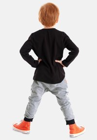Musta pitkähihaine paita, harmaat jogger-housut ja oranssit korkeat tennarit. Lapsi ottaa itsevarman asennon kädet lanteilla.