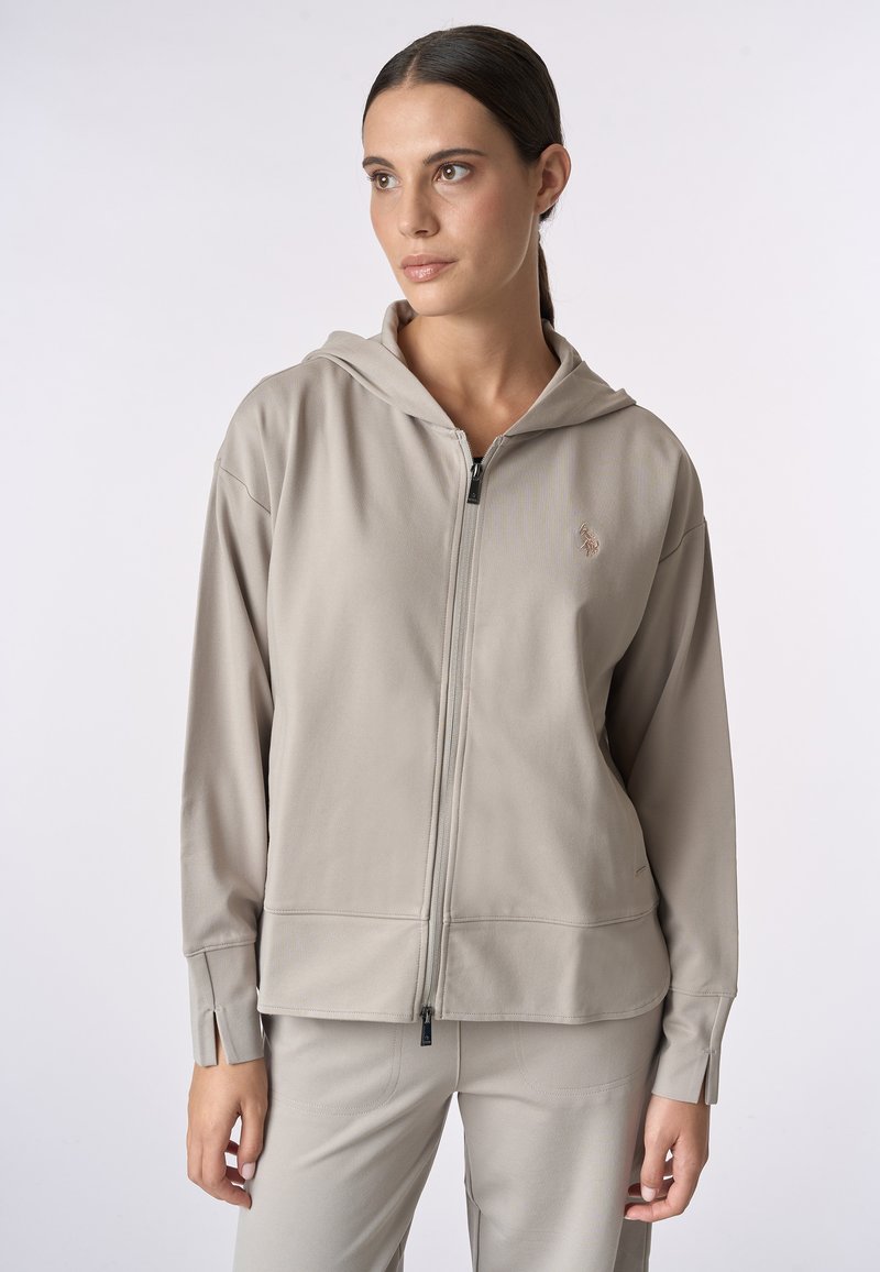 Felpa con zip di colore beige, realizzata in tessuto morbido con cappuccio e una sottile texture. Presenta una zip frontale e un logo ricamato sul petto.