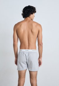 Jaunas vyras su garbanotais tamsiais plaukais stovi be marškinėlių, atsisukęs nuo kamerų, vilkintis šviesiai pilkus dryžuotus Calvin Klein apatinukus prieš paprastą foną.