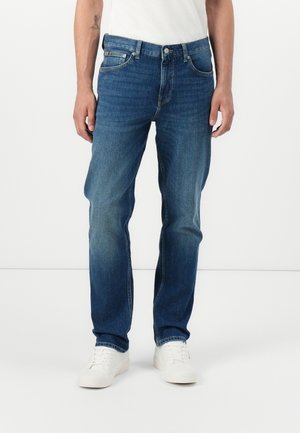 Mężczyzna ubrany w niebieskie, proste dżinsy o średnim odcieniu z lekko wyblakłym efektem, białe trampki i białą koszulkę, stojący na tle gładkiego, białego tła.