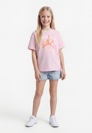 Mlaka djevojka s dugom plavom kosom nosi ružičastu majicu s grafičkim uzorkom, svijetloplave traper kratke hlače i bijele tenisice, smiješi se u kameru.