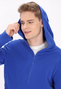 Felpa blu in pile con zip frontale e cappuccio foderato. Il tessuto sembra morbido e strutturato, con un design semplice e rilassato.
