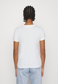 Person with braided hair wearing a plain white short-sleeve T-shirt and light blue jeans, facing away against a neutral background.