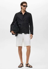 Camisa negra de botones hecha de un material texturizado, pantalones cortos blancos, sandalias negras y gafas de sol. Modelo sosteniendo una chaqueta negra.