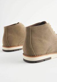 Stivaletti ankle in suede marrone con suole in gomma bianche e marroni, caratterizzati da cuciture dritte e tacco basso. Vista laterale che evidenzia la texture e il design.