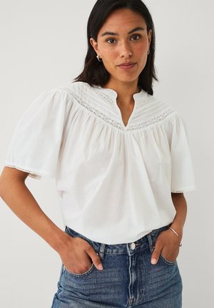 Kvinde iført en hvid bluse med pufærmer og blondekant og blå jeans, stående med hænderne i lommerne mod en ensfarvet baggrund.