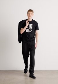 Svart kortermet t-skjorte med grafisk design, svarte bukser og svarte sko. Modell som holder en svart blazer, står mot en ensfarget bakgrunn.