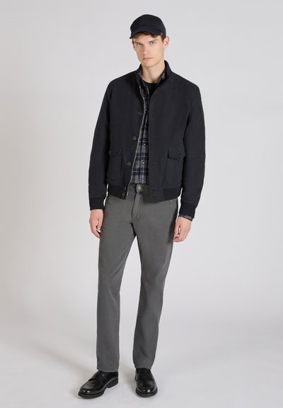 Marine blauwe jas met knoopsluiting en twee voorzakken, gelaagd over een geruit shirt, gecombineerd met grijze broek en zwarte schoenen, afgewerkt met een pet.