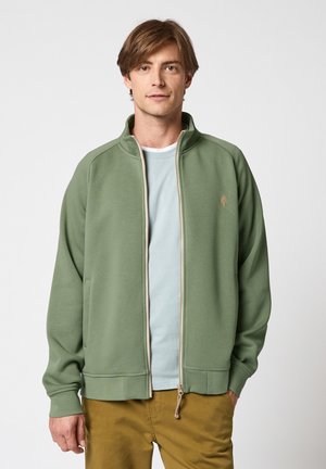 Jeune homme portant une veste zippée vert clair sur une chemise bleu pâle et un pantalon moutarde, debout devant un fond blanc uni.