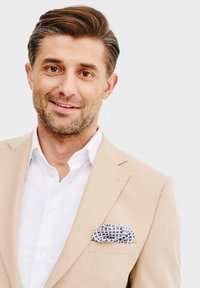 Beżowy lniany marynark z białą koszulą pod spodem, z wzorzystą kieszonkową chusteczką w błękicie i bieli. Neutralne tło.