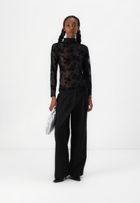 Čierny kvetinový čipkový top s dlhým rukávom v kombinácii s voľnými čiernymi nohavicami. Modelka drží strieborný saténový clutch a má na sebe nadmerné kruhové náušnice.